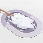 Purple-grey oval bath mat seen from above with a hand pressing soapy foam onto its surface
