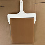 Wooden sliding pizza peel with brown fabric belt and long handle, photographed on cardboard
